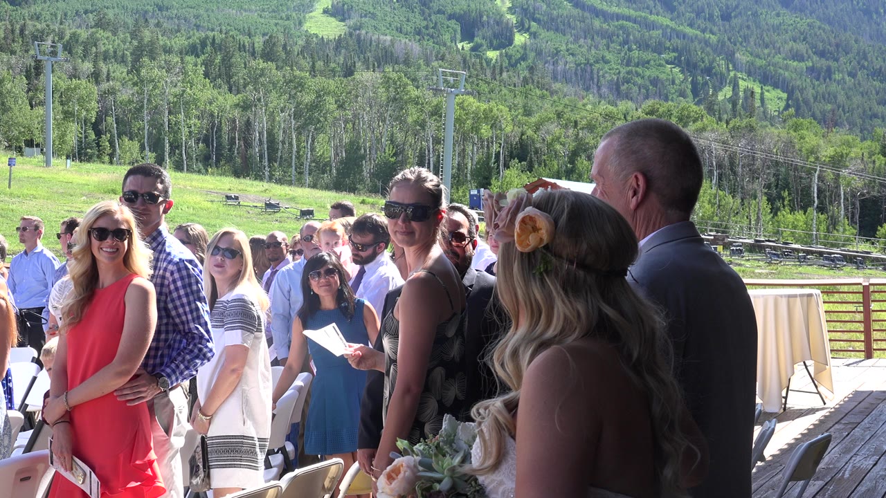 Bride and father approach outdoor wedding guests with green hills behind them.