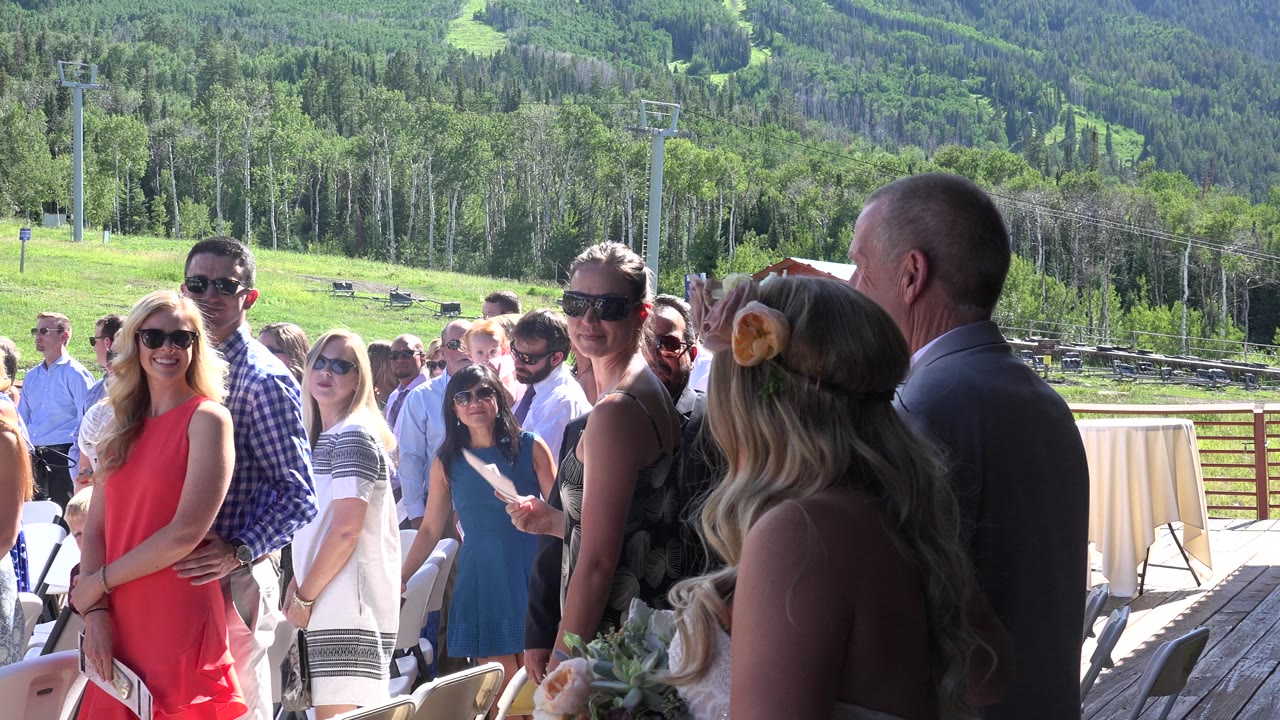 The bride and the man in the gray suit continue down the aisle while smiling guests in summer clothing watch from both sides under bright sun in Mesa, Colorado.