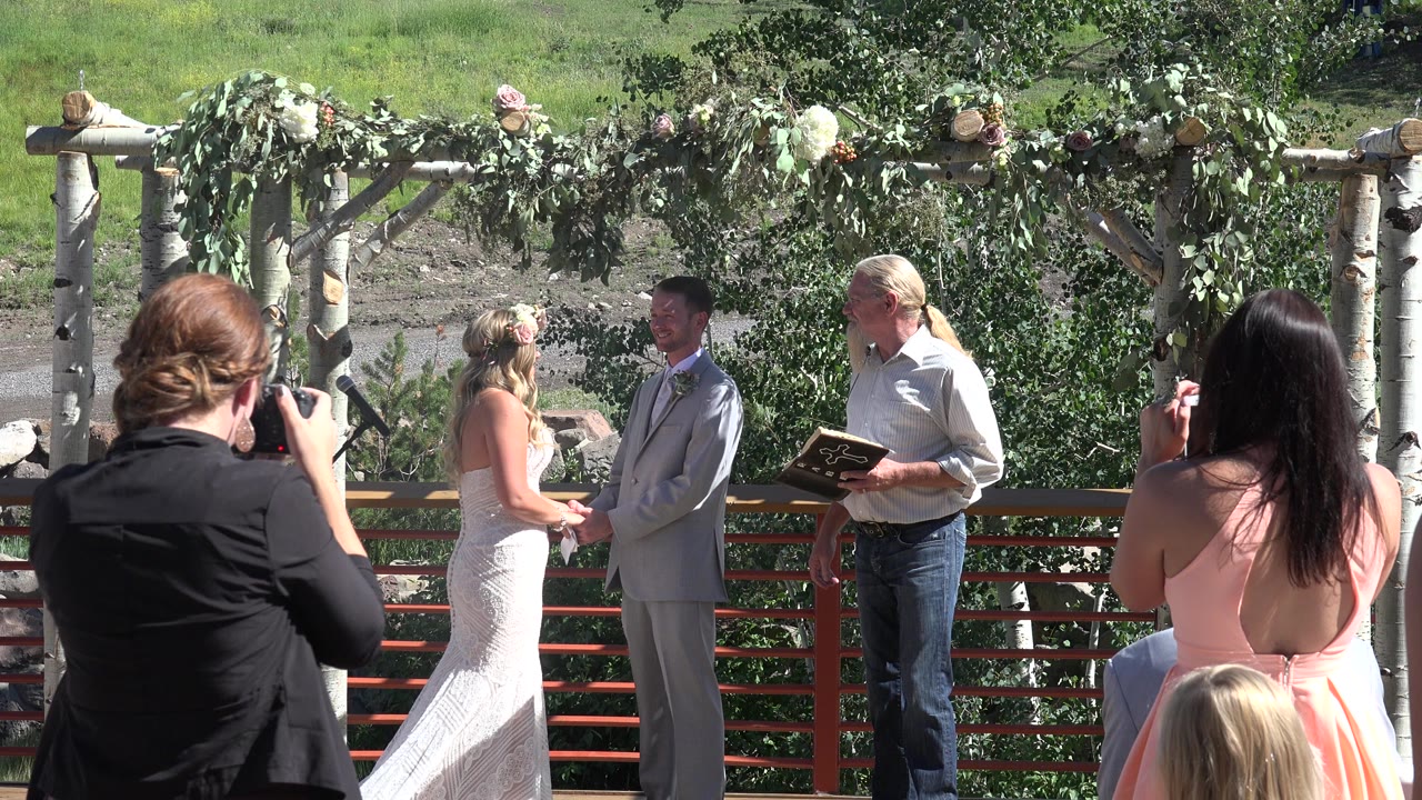 In Mesa, Colorado, a bride in white and a groom in a light gray suit face each other under a leafy wedding arch while an officiant holds a book and guests watch from the foreground.