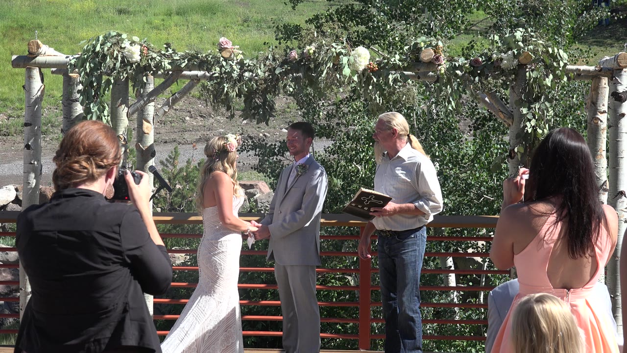 In Mesa, Colorado, the bride and groom remain under the branch arch as a foreground guest raises a camera and the officiant stands to the side with an open book.