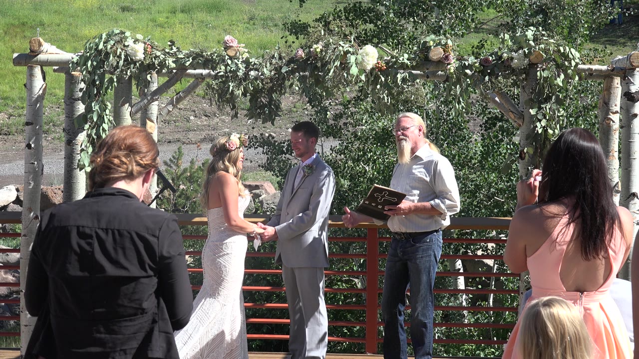 Officiant smiles beside a bride and groom during an outdoor ceremony.