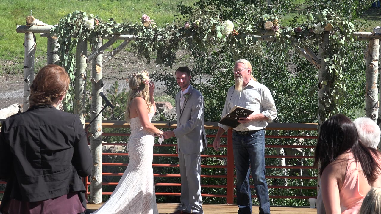 Bride and groom stand beneath a leafy wedding arbor with an officiant nearby.