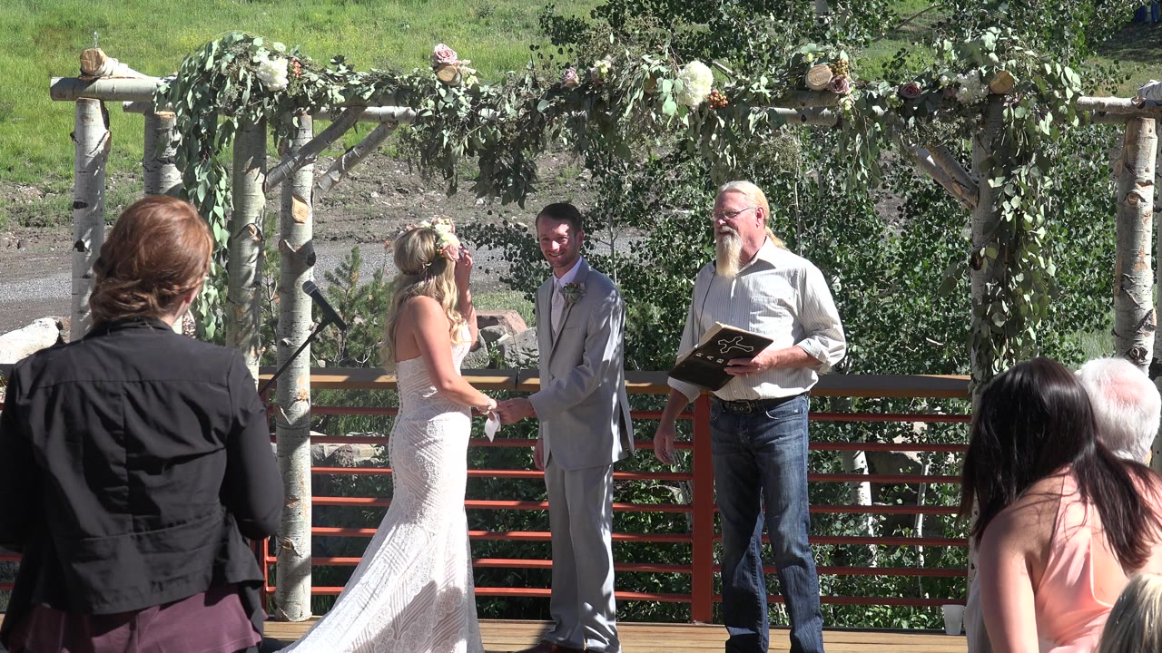 At 0:01 in Mesa, Colorado, the bride and groom hold hands under the decorated arbor, with the officiant looking on and seated guests partly blocking the lower frame.