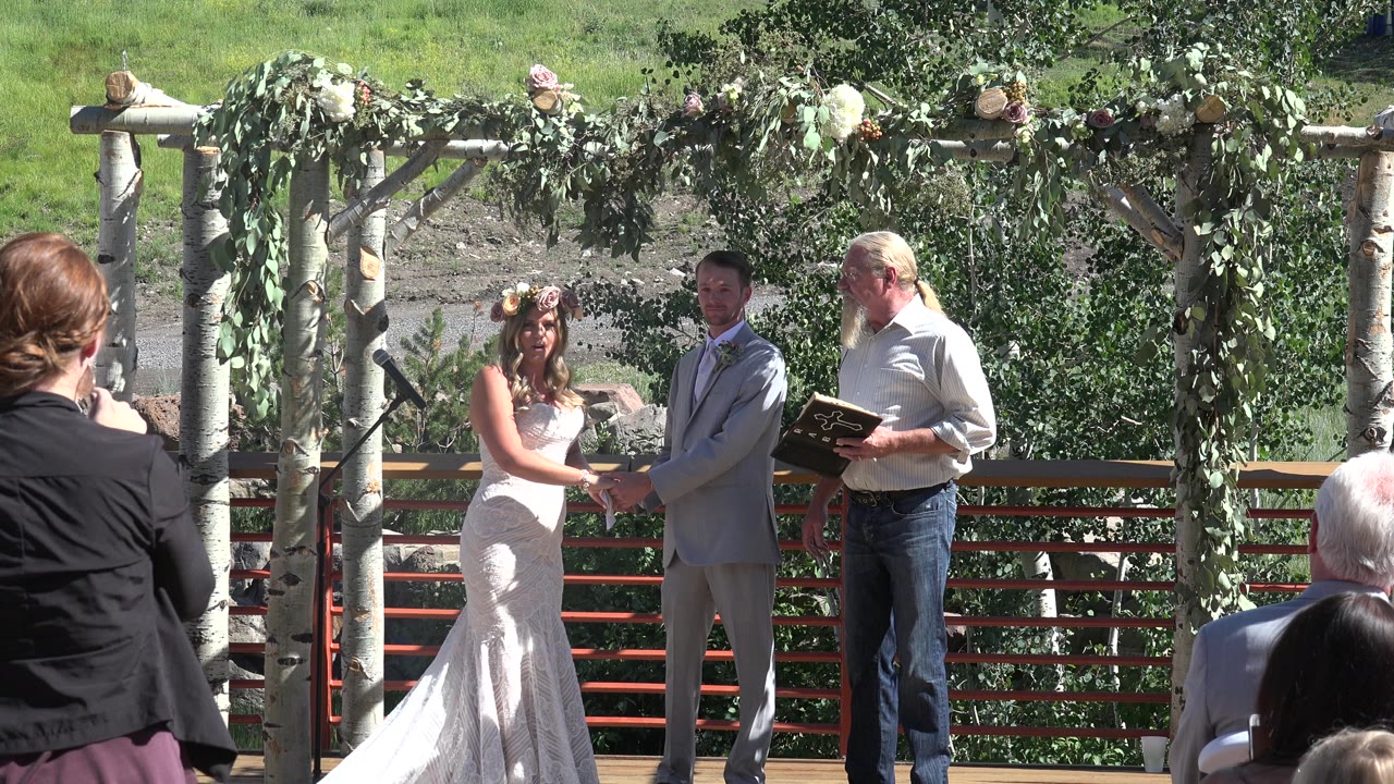 Bride and groom holding hands under a leafy arch with an officiant nearby.