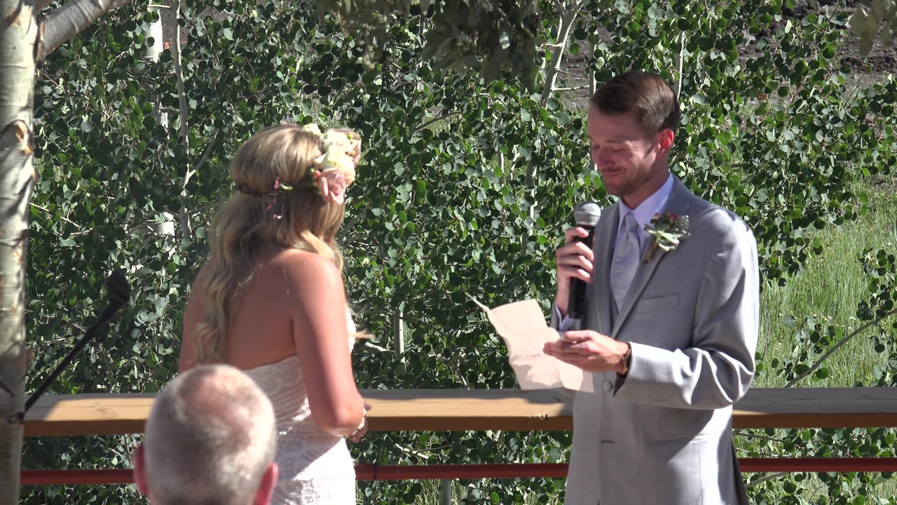 One second in, the bride remains turned toward the groom as he reads from a sheet beside a railing, with bright green trees behind the ceremony space in Mesa, Colorado.