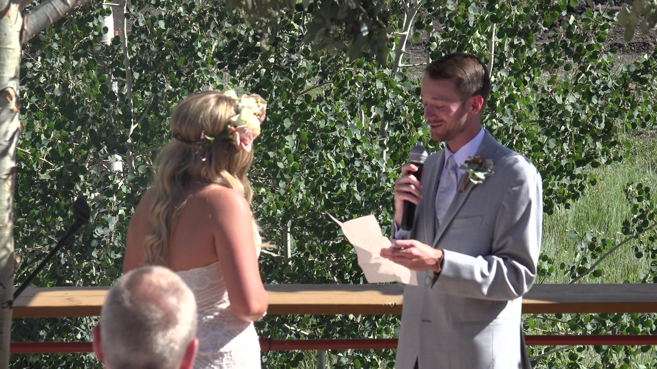 Groom smiles while reading from paper at outdoor ceremony.