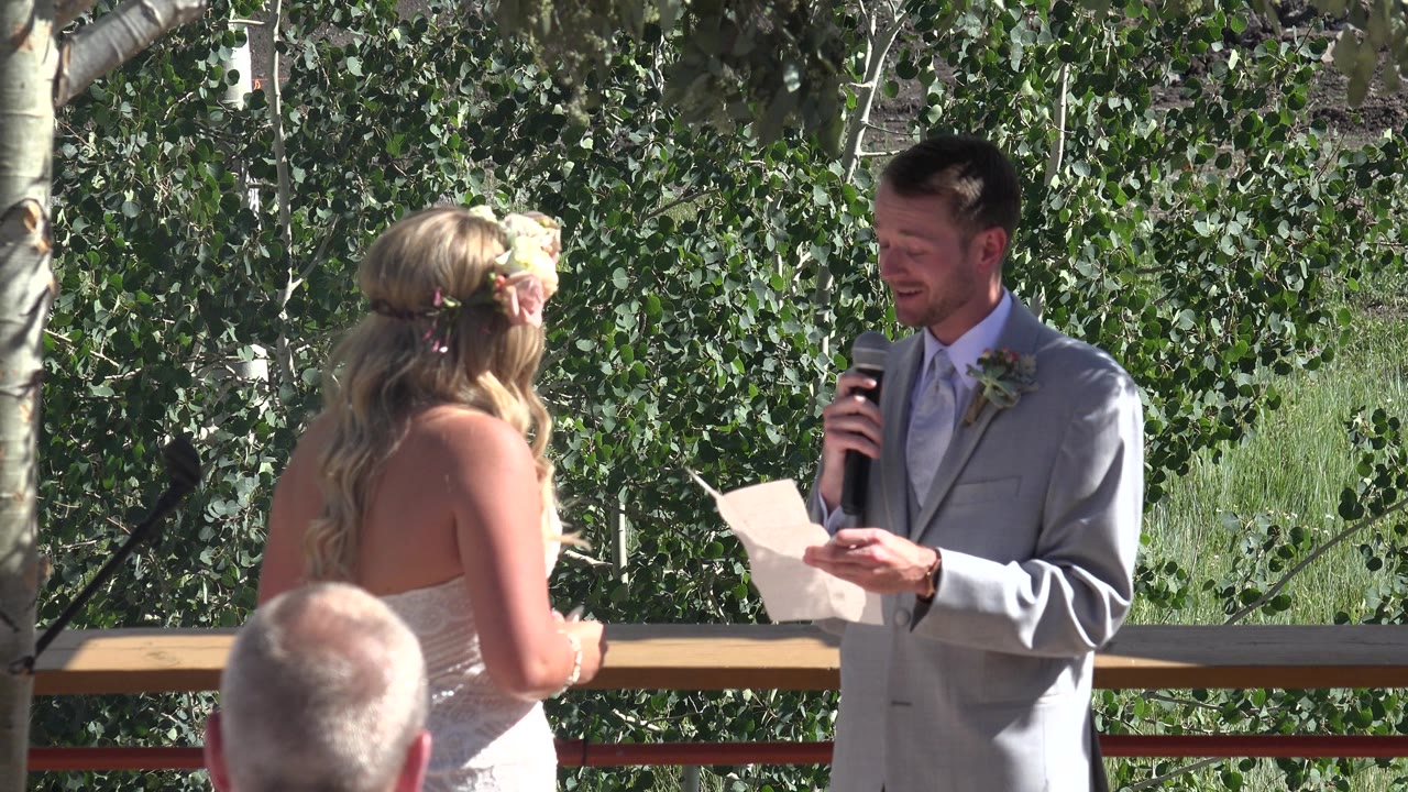 Bride facing groom as he reads into a microphone outdoors.
