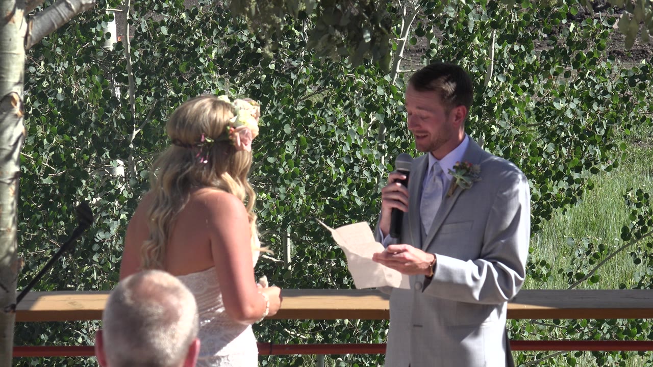 One second in, the groom continues facing the bride, holding a microphone near his mouth and notes in his other hand against leafy Mesa, Colorado trees.