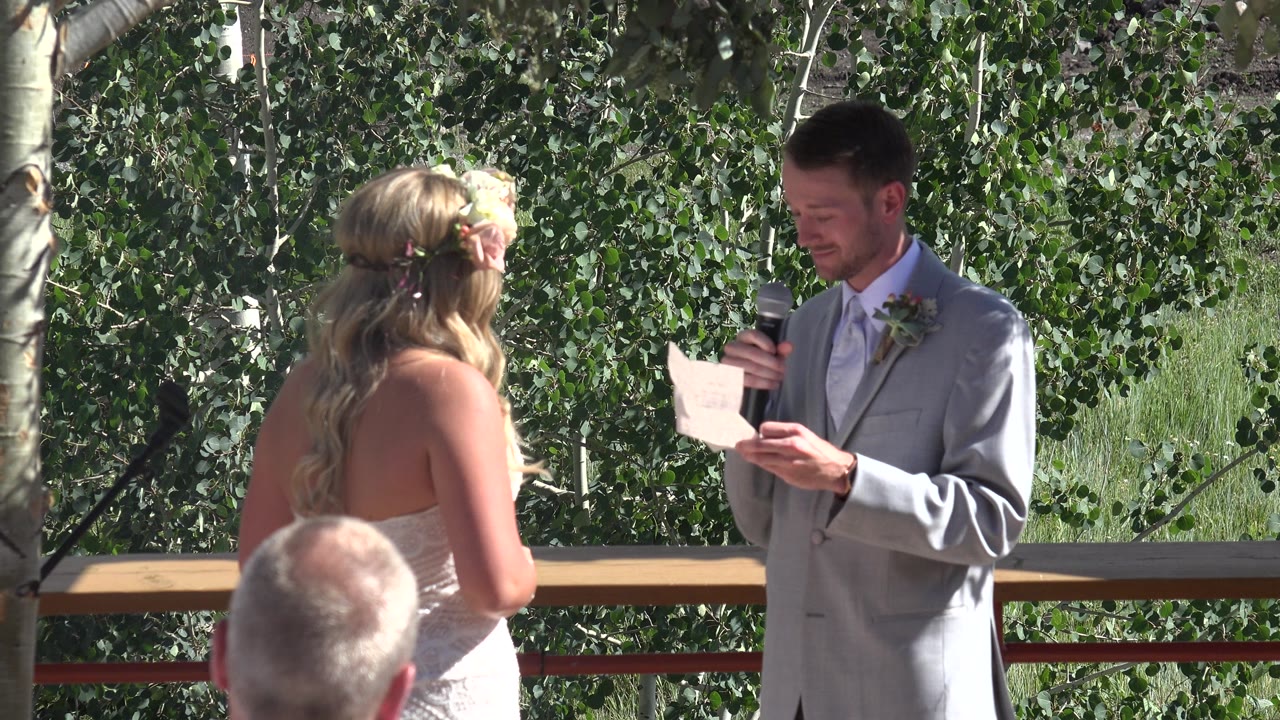 At 0:01 in Mesa, Colorado, the groom looks down at his paper with the microphone raised, while the bride stands opposite him near a railing and dense green trees.