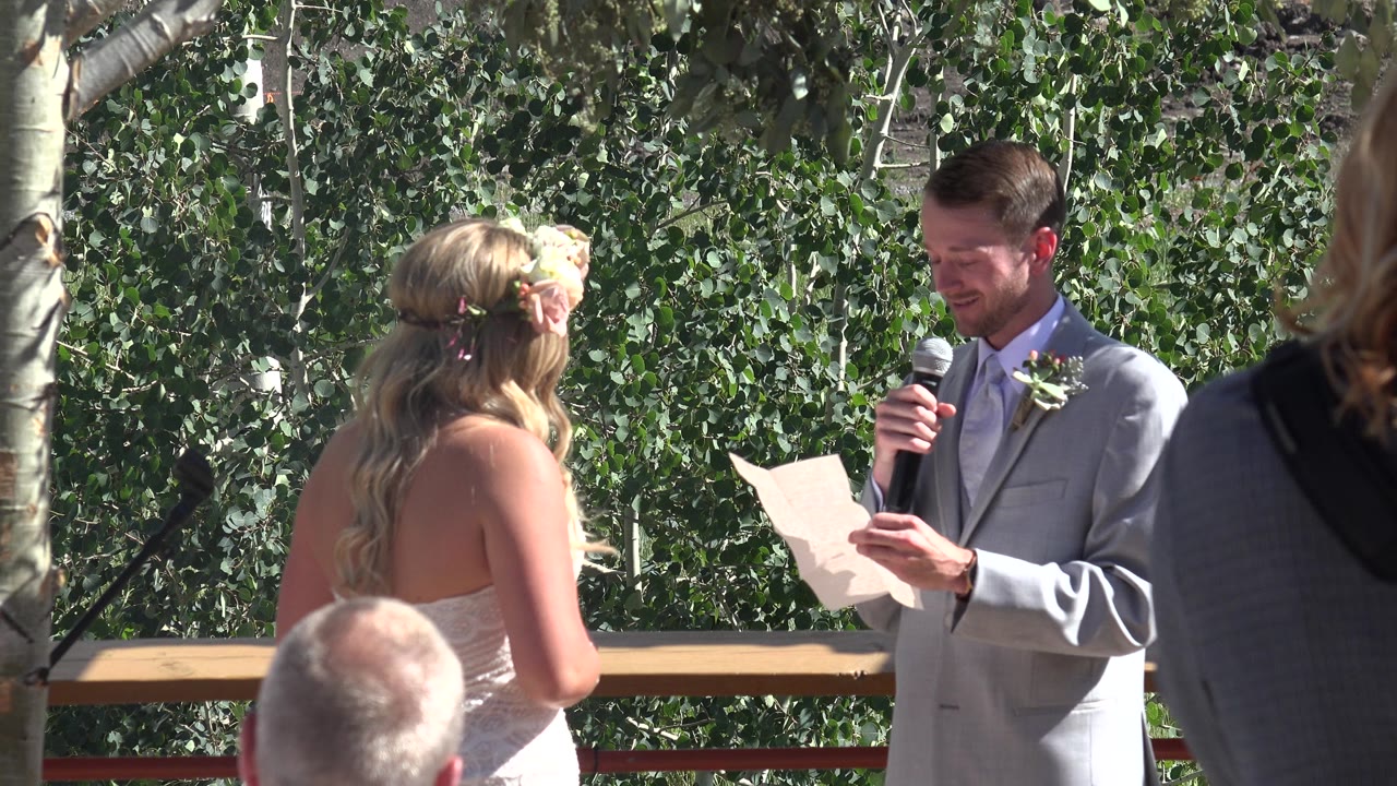 Bride faces a suited man reading from paper with a microphone outdoors.