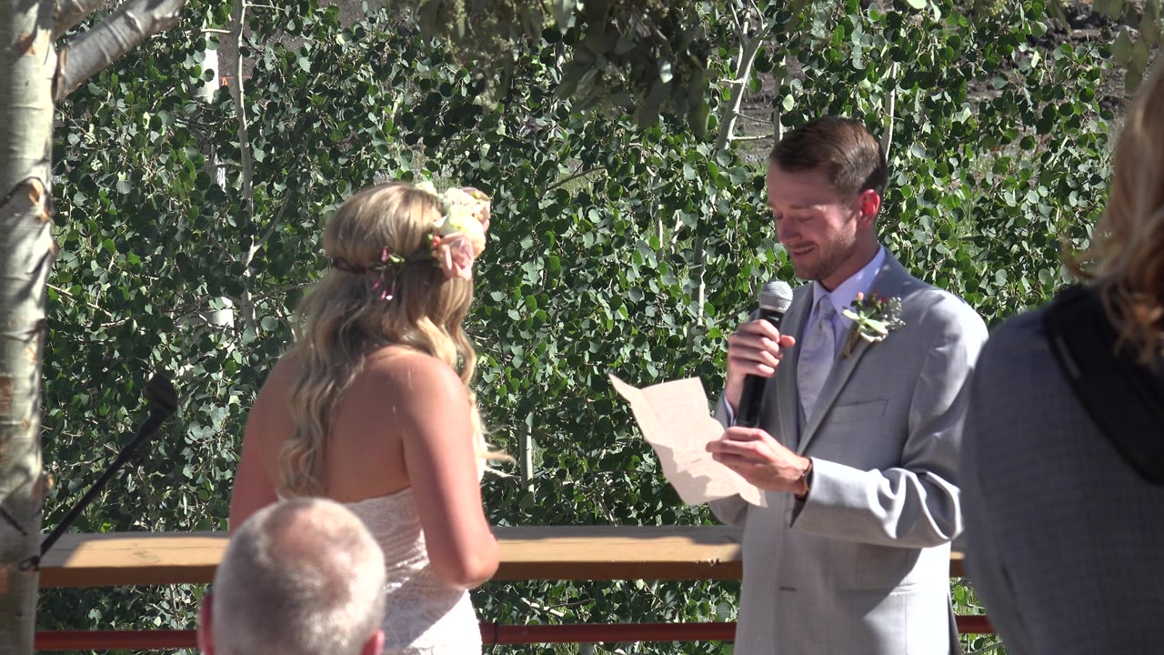 At 0:01 in Mesa, Colorado, the suited man looks down at his paper while the bride stands opposite him and leafy trees fill the sunlit background.