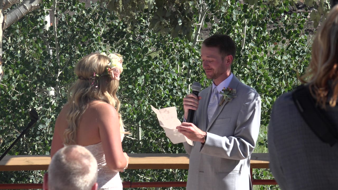 In Mesa, Colorado, a man in a light grey suit reads from a white paper while holding a microphone toward a blonde woman in a white dress, with guests and trees behind them.
