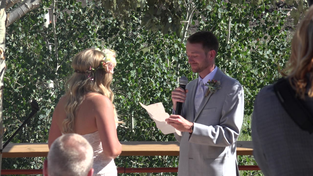 Bride faces man reading from paper in front of trees.