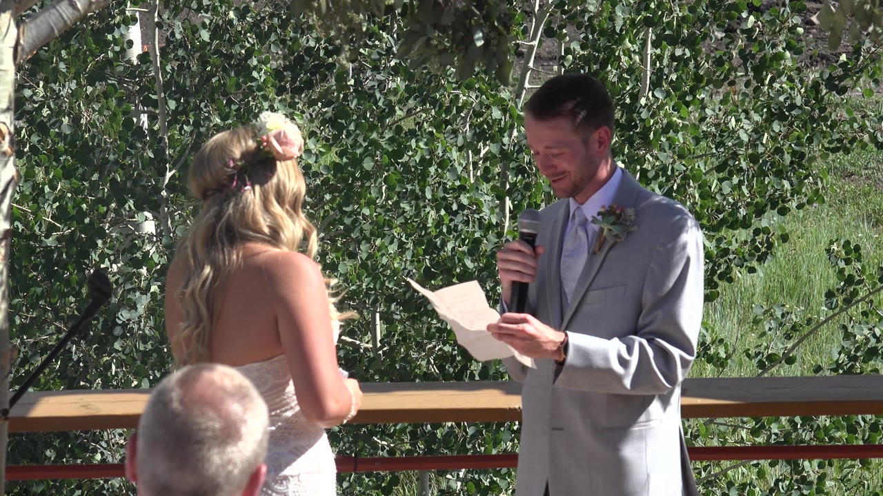 In Mesa, Colorado, a bride seen from behind faces a groom in a light gray suit as he reads from paper into a microphone beside leafy trees.