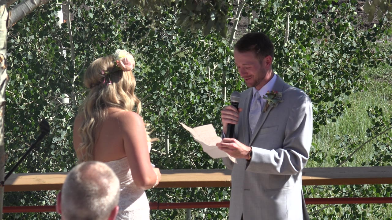 The groom stands on the right holding a microphone and paper while the bride listens with her back to camera in the Mesa, Colorado outdoor wedding setting.