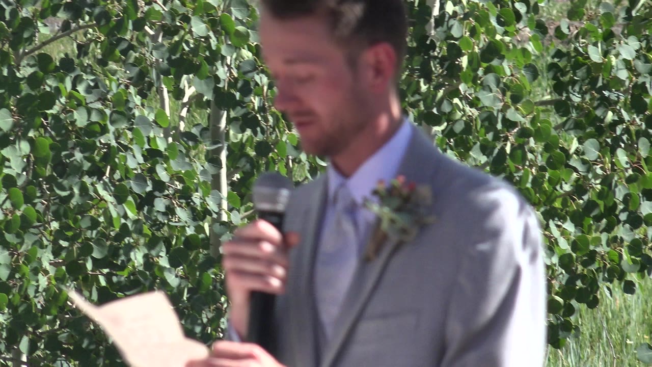 At 0:00 in Mesa, Colorado, a groom in a gray suit holds a microphone while a sheet of paper appears in the lower foreground against green trees.