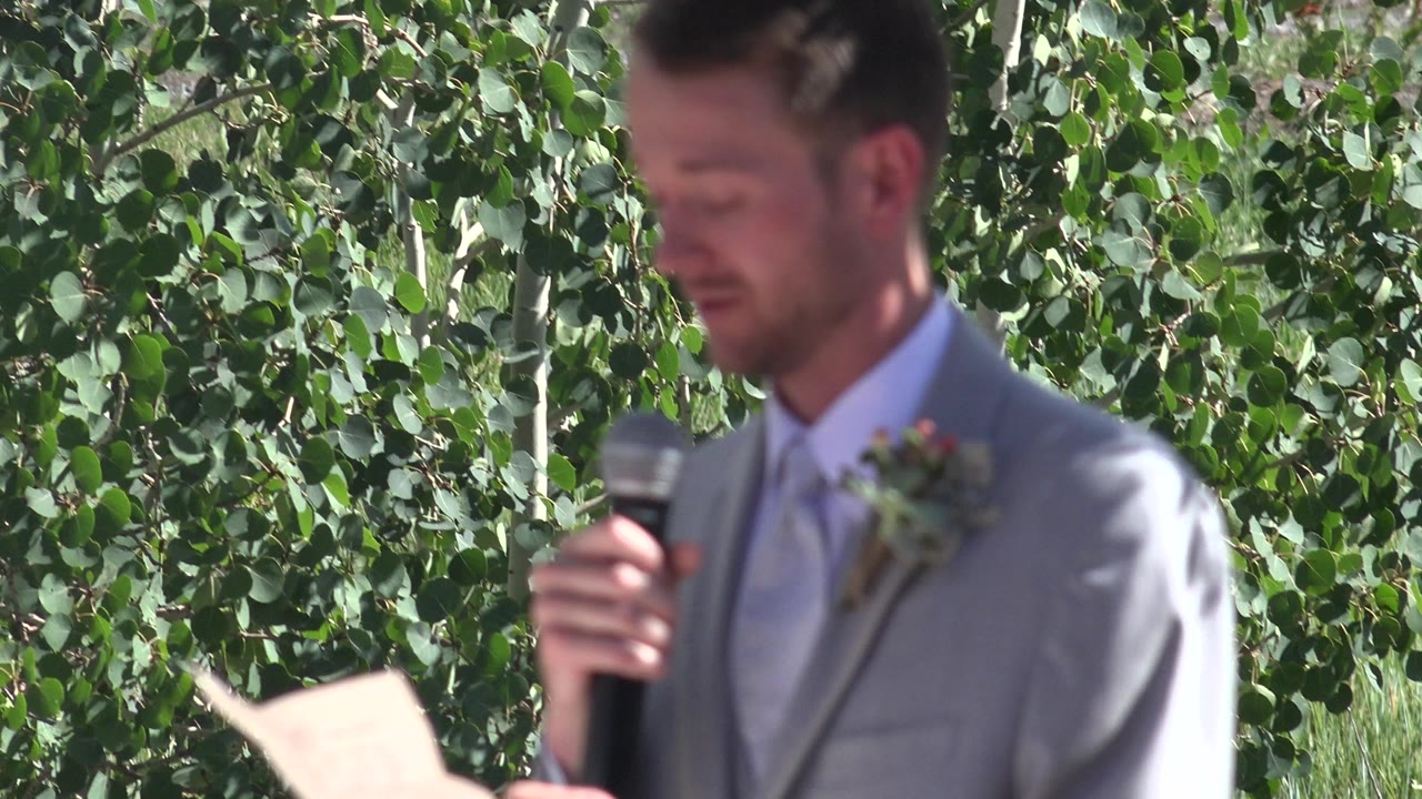 At 0:01 in Mesa, Colorado, the groom looks down with the microphone near his mouth as a hand-held paper remains at the lower edge of the frame.