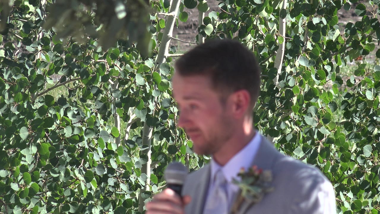 Groom in suit holds a microphone in front of leafy trees.