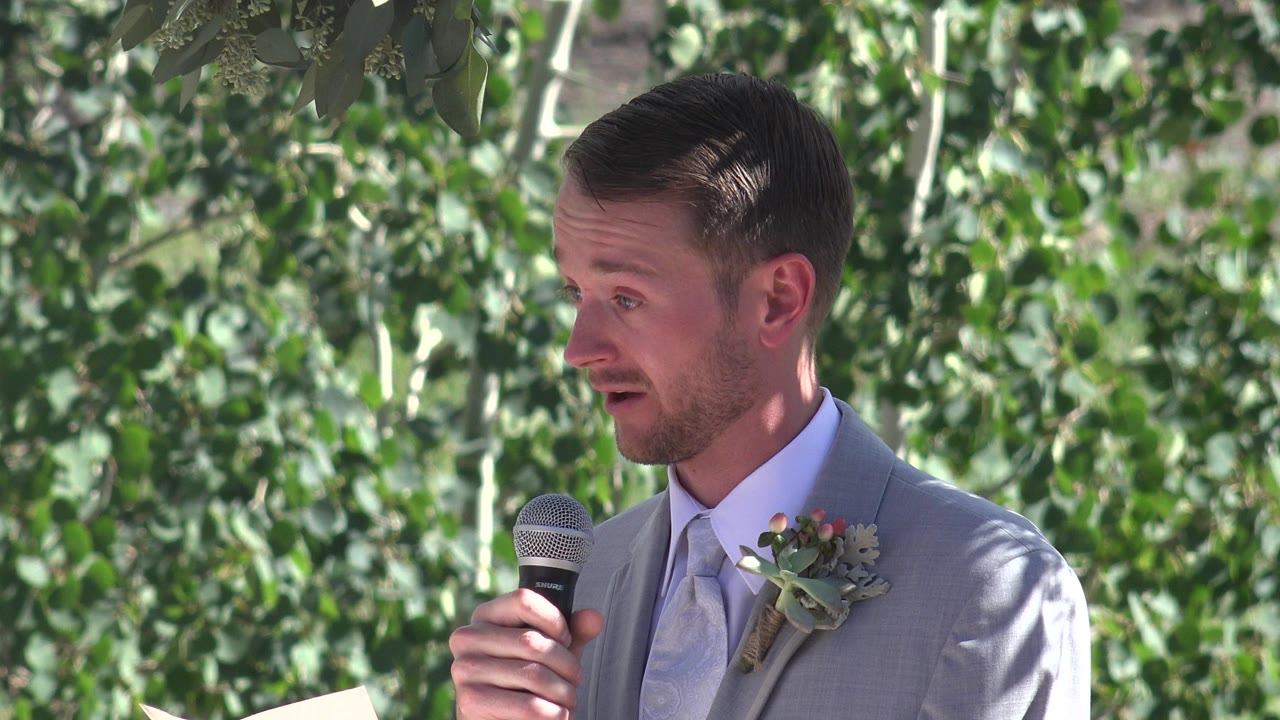 Man in a gray suit holds a microphone outdoors beside green leaves.