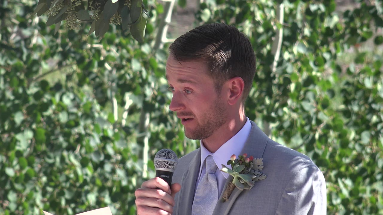 At 0:01, the suited speaker holds the microphone near his mouth in the Mesa, Colorado outdoor setting, with dense green leaves filling the background.