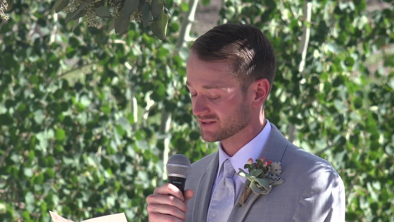 Groom in a gray suit holds a microphone outdoors.