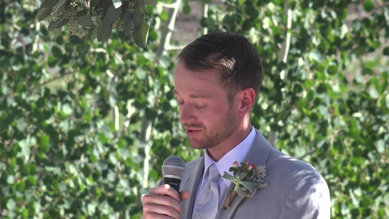Groom in a gray suit holding a microphone before leafy trees.
