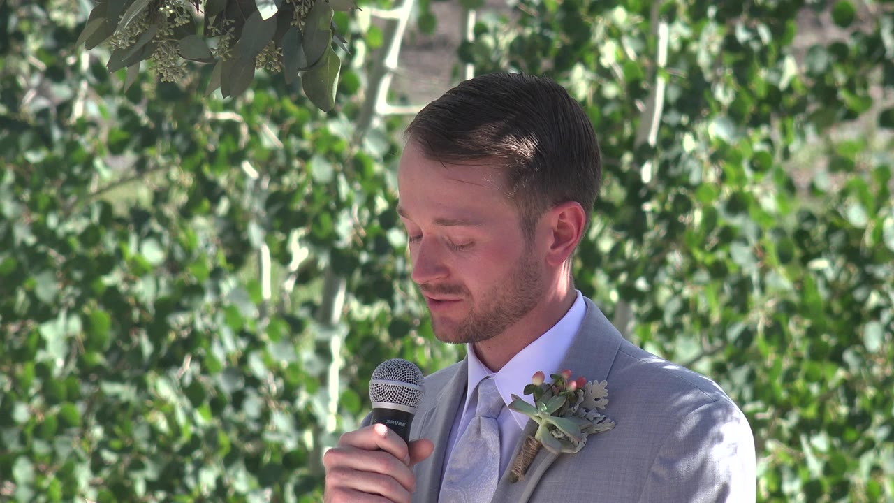 The groom looks downward while holding the microphone, with his boutonniere visible on a light gray jacket and sunlit leaves filling the Mesa, Colorado background.