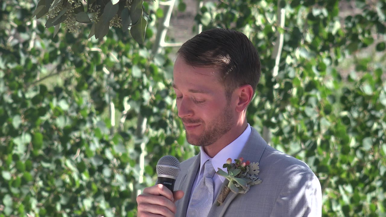 Man in gray wedding suit holding a microphone under leafy trees.
