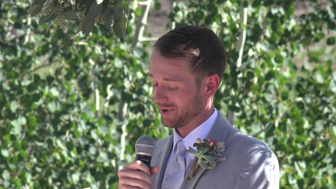 In Mesa, Colorado, the suited man keeps his eyes lowered and holds the microphone closer as leafy branches fill the background.