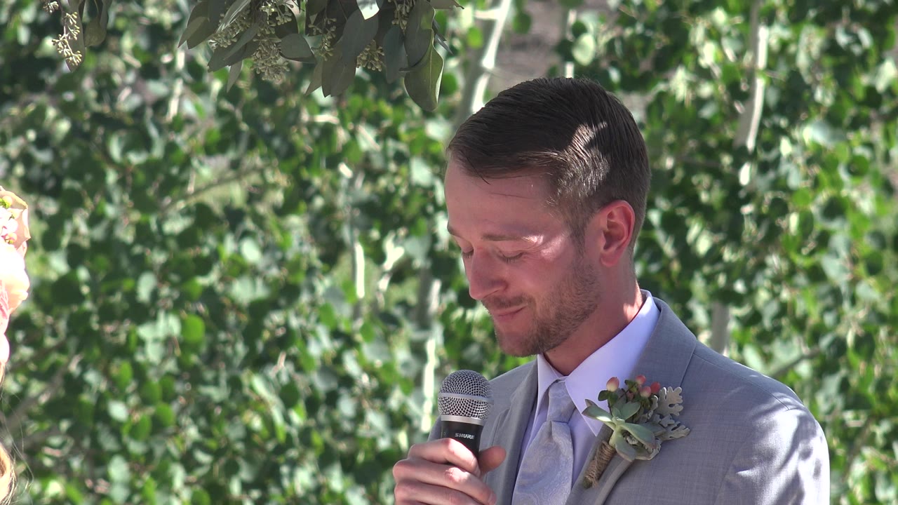 At 0:00 in Mesa, Colorado, a groom in a light gray suit looks down while holding a microphone in front of leafy green trees.