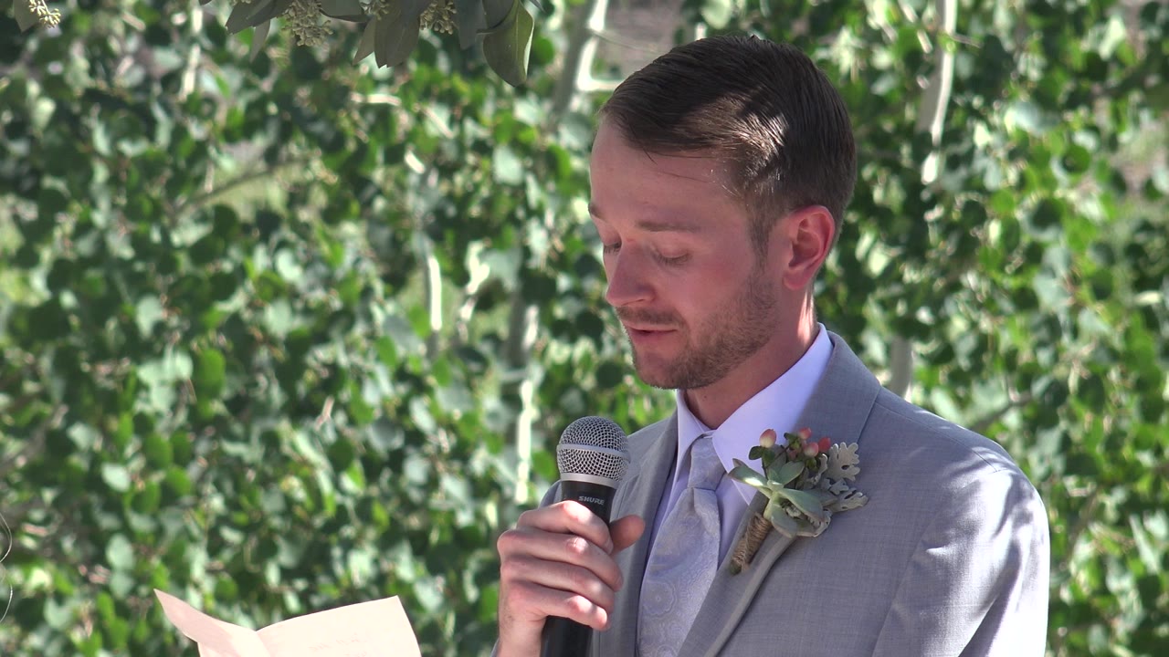 Man in gray suit holds a microphone while reading from paper outdoors.