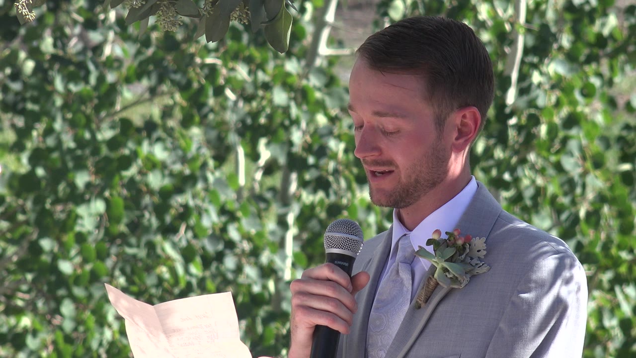 Groom in a gray suit reads vows into a microphone outdoors.