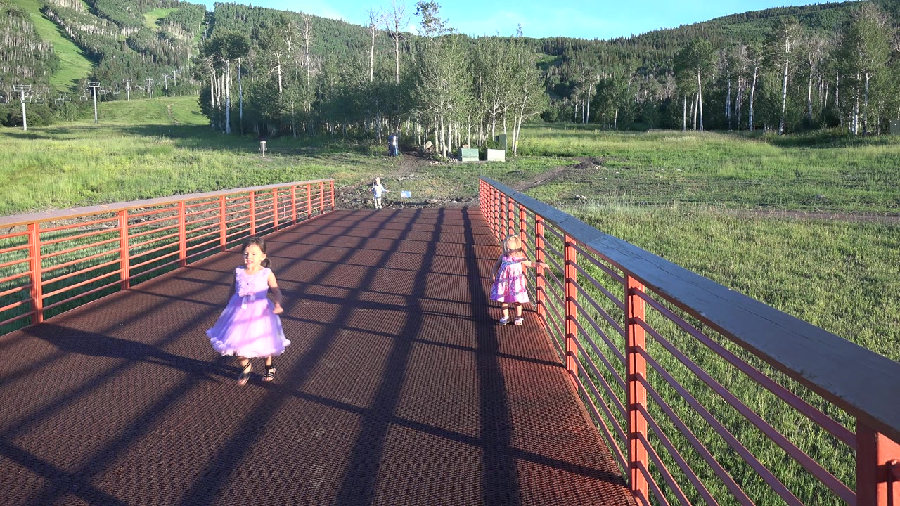 A child in a purple dress moves closer across the sunlit red bridge in Mesa, Colorado, while another child stands near the right railing and grassy hills fill the background.