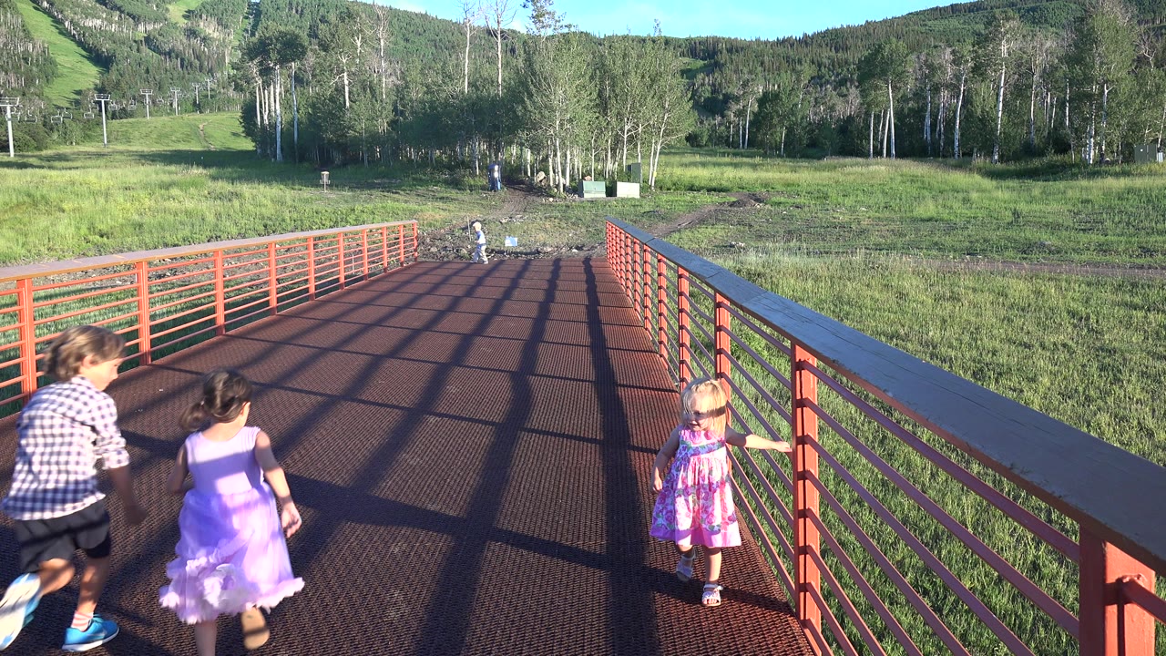 Children running onto a red metal bridge near grass and trees.