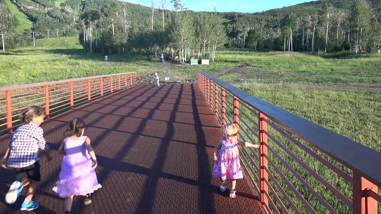 Children continue across the red bridge in Mesa, Colorado, while rail shadows stretch over the walkway and a grassy slope fills the background.