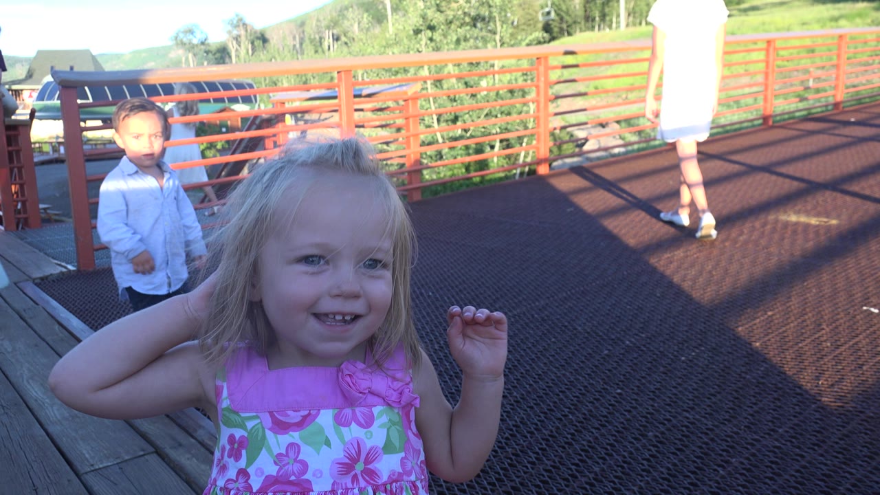 The blonde girl remains close to camera on the Mesa bridge, smiling beside red railings while a boy and adults stand farther along the sunlit deck.