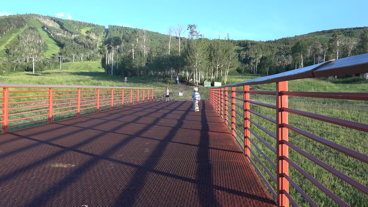 In Mesa, Colorado, a red-railed bridge leads toward a green hillside while three children are visible farther down the walkway.