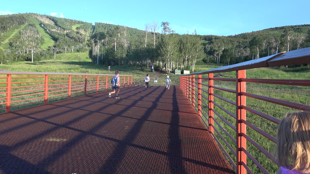 At 0:01, the children are farther along the red walkway in Mesa, Colorado, while a blond child's head appears at the lower right foreground and the hillside remains in view.