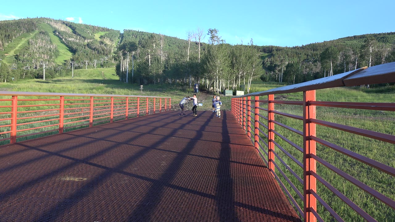 Children stand and run on a red metal bridge in Mesa, Colorado, with green grass, trees, ski-lift towers, and wooded hills in the background.