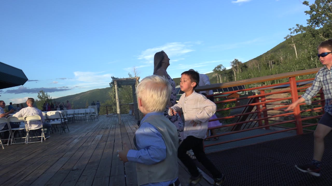In Mesa, Colorado, children move across a wooden deck while adults sit and stand near white folding chairs, with a red railing, hills, and blue sky in the background.