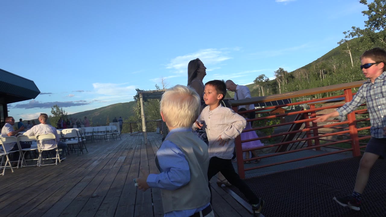 In Mesa, Colorado, two children run near a red railing as adults and white folding chairs remain visible on the wooden deck beneath a clear blue sky.