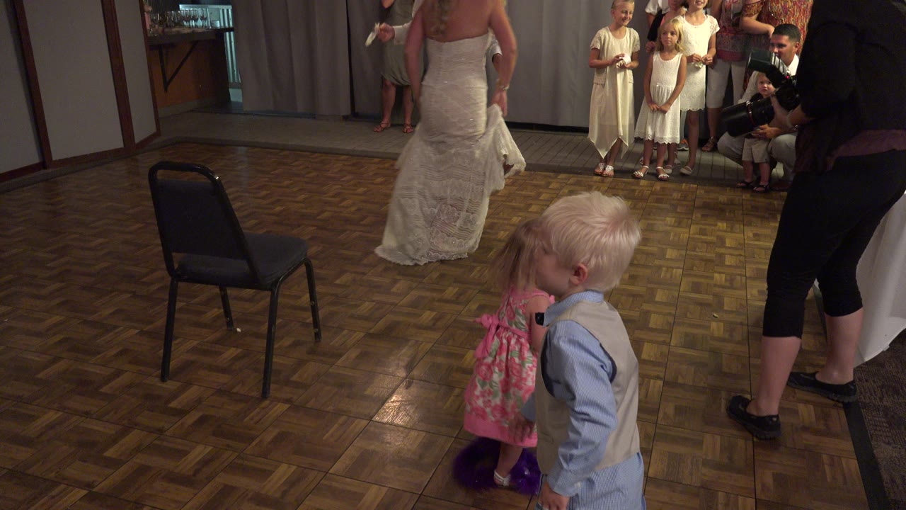 At a Mesa, Colorado indoor gathering, a blond boy and a little girl stand in the foreground while adults and children watch from near the back wall.