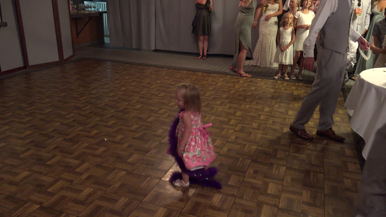 In Mesa, Colorado, a little girl in a pink floral dress kneels on a parquet dance floor with a purple boa while adults and children stand near curtains in the background.