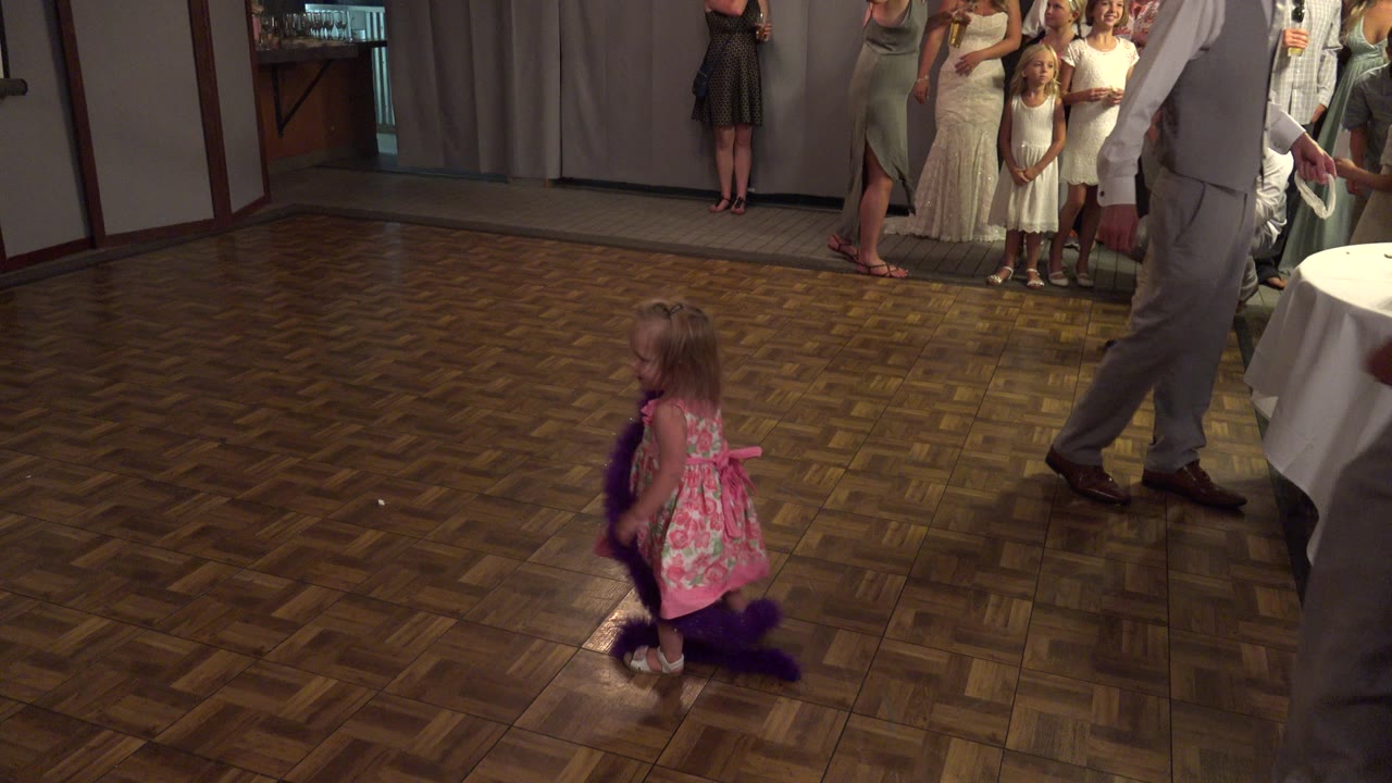 A small girl crouches with a purple boa on the parquet floor as an adult in a gray suit crosses the right side of the Mesa party room.