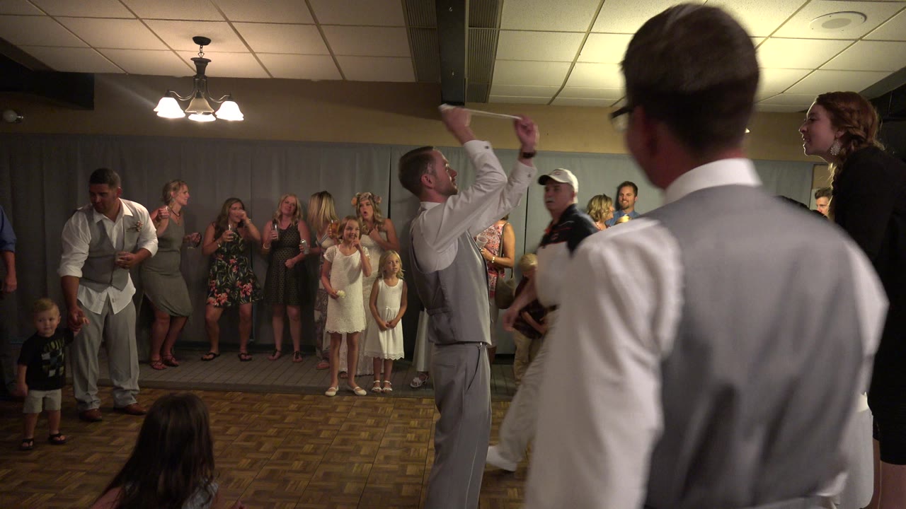 At a Mesa, Colorado wedding reception, guests and children line the back wall while men in light vests and suits stand on the dance floor under warm ceiling lights.