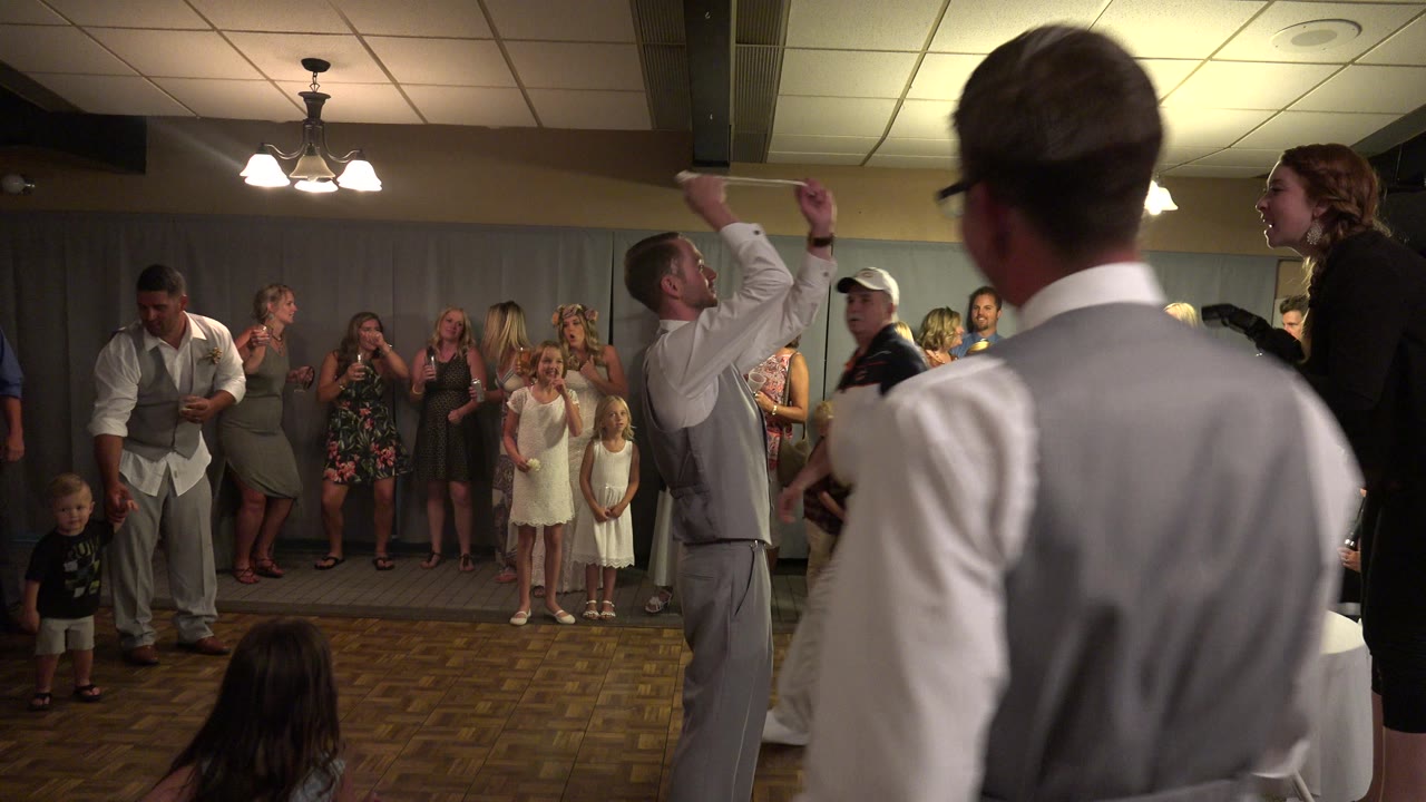 A man in a gray vest raises his arm near the center of the dance floor while a man in glasses is close to camera and wedding guests watch from the curtained wall in Mesa, Colorado.