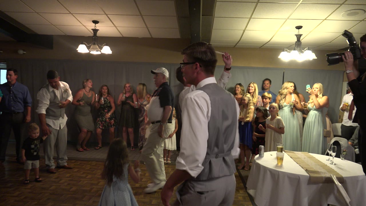 Wedding party and guests stand near a banquet table under bright indoor lights.