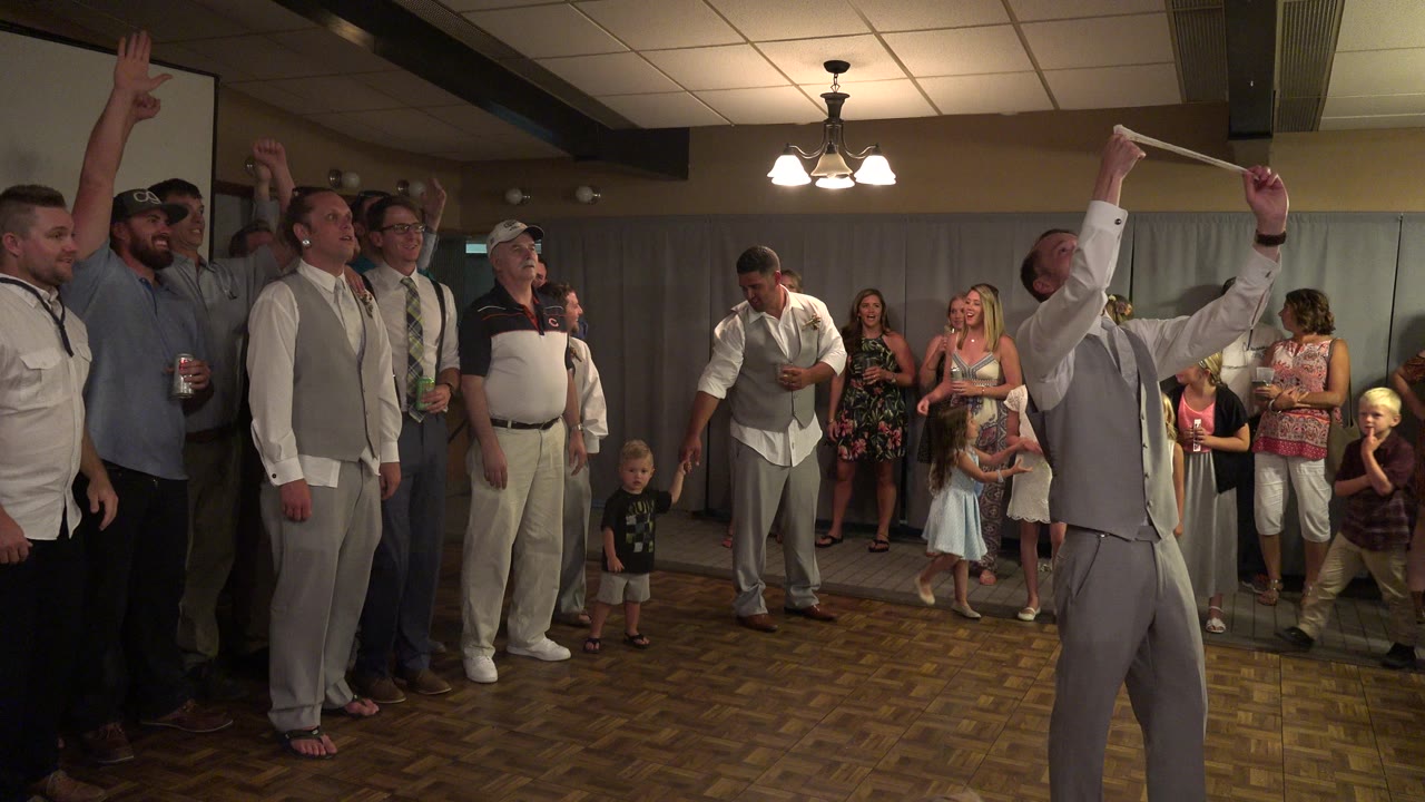 At a Mesa, Colorado wedding reception, several men in dress shirts and vests stand on a parquet dance floor with arms raised while guests and children watch from the back of the room.