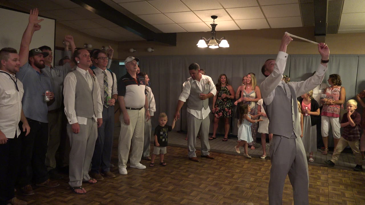Wedding guests in Mesa, Colorado cluster around the dance floor as a man in a gray vest raises both arms and adults and children stand along the curtained wall.