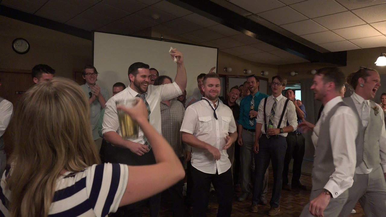 A smiling group of adults in Mesa, Colorado fills the party room while a man in a tie raises one hand and another man in suspenders stands nearby.
